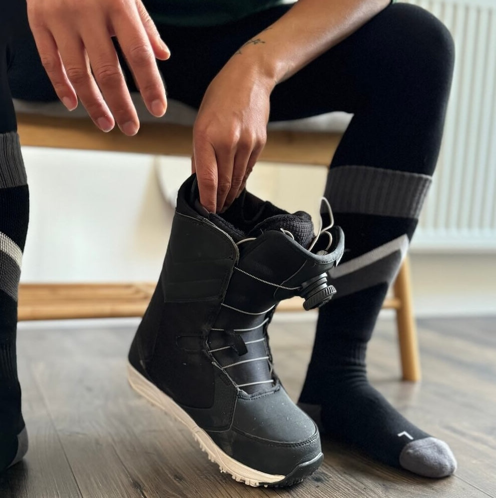 Close-up of a person sitting on a wooden bench, wearing black and grey Do Better Colab organic cotton ski socks with a diagonal stripe, holding a black snowboard boot on a wooden floor.