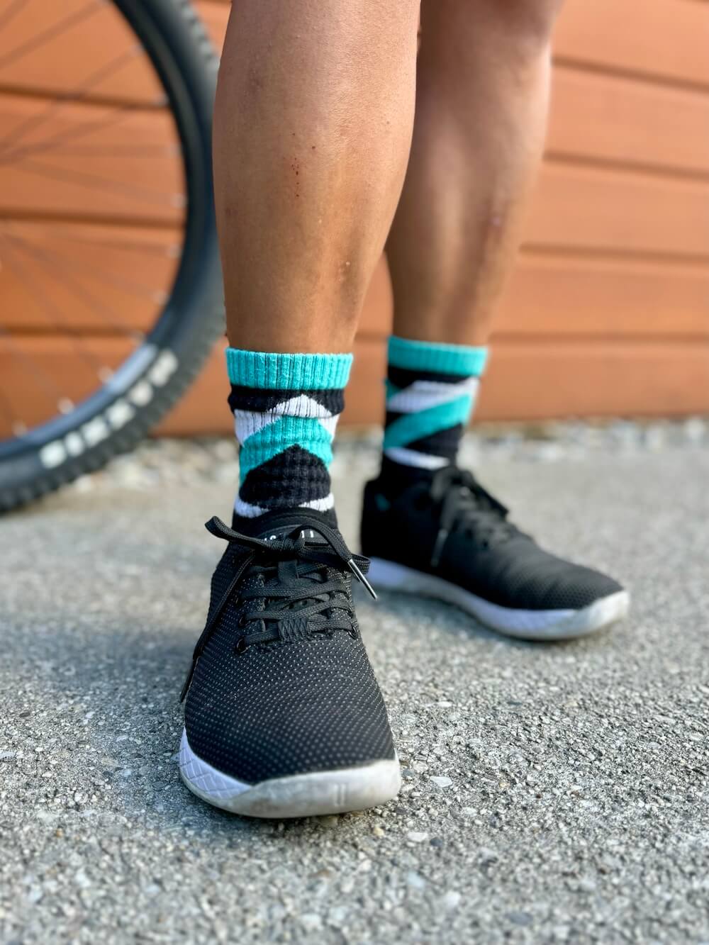 Close-up of a person wearing black trainers and teal, black, and white Do Better Colab organic cotton crew socks with a chevron pattern, standing on a gravel path next to a mountain bike tyre.