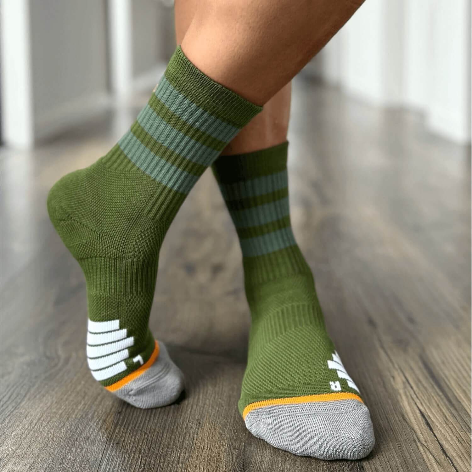 Close-up of a person wearing green Do Better Colab organic cotton crew socks with lighter green stripes and grey toes featuring white and orange detailing, standing on a wooden floor.